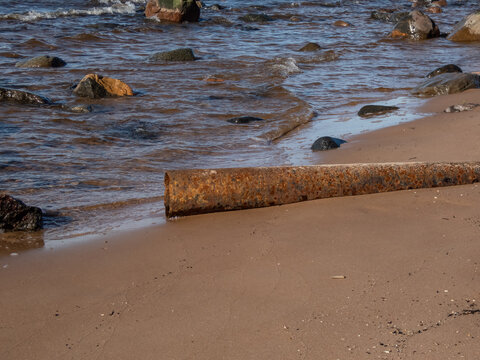 Big metallic pipe covere with orange rust on a beach of sea shore partly in water surrounded with big rocks