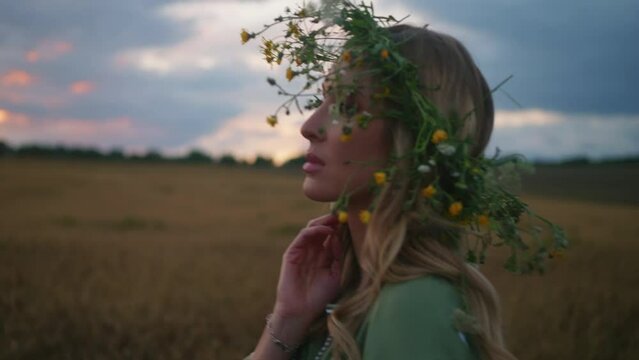 Young woman with floral crown walks thoughtfully at wheat field. Feeling hopefull, wishing of peace. Bright make up, embroidered green dress. Colorful sunset out of the city. High quality 4k