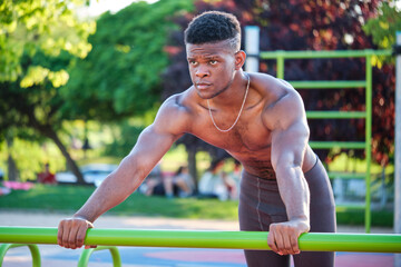 Young fit shirtless black man doing push-ups on a bar in a calisthenics park outdoors on sunny day....