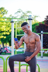 Obraz premium Portrait of a young fit shirtless black man smiling and using the smartphone in a calisthenics park outdoors. Fitness and sport lifestyle.