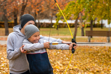 Asian laughing 7 years boy learns to shoot an arrow with the help of his older brother outdoor in public park in autumn, close up, selective focus.