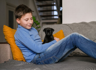 Boy playing with baby dog. Kid play with puppy at home. Little boy and griffon or brabanson dog on sofa. Children and pets at home. Kid sitting on the floor with pet. Animal care.