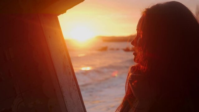 Woman stands in old wooden ruined leaning house interior on sea beach, looks at window on sunset. Mixed ethnic female enjoys view from vintage abandoned rickety home on coastline. Dreamy, enigmatic.