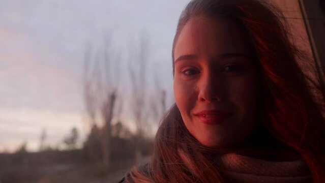 Enigmatic woman sits on windowsill of abandoned wooden leaning house on sea beach, looks at window on sunset, on camera, smiles. Dreamy girl enjoys view in vintage rickety home. Wind blows airy hair.