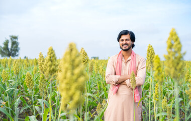 Confident young indian farmer standing with crossed arms by looking camera at farmland - concept of...