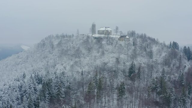 Aerial shot of a church on top of a mountain with a snowy forest in winter, in Slovenia