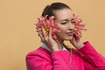 Fototapeta premium Smiling brunette woman in pink sweater wearing floral headphones and listening music. Dahlia flower buds as a earphones.