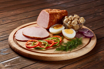 Beautifully presented dish of sliced brisket, jamon and bacon with bread, vegetables, herbs, eggs, sauce on a round wooden board on a dark wooden table