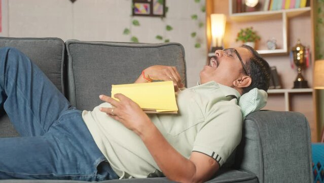 Middle aged man falling sleep while reading book or novel on sofa at home - concept of boredom, fatigue and relaxed.