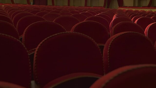 Antique armchairs upholstered in soft red fabric. Rows of chair backs in an empty opera house. Bright soffit light.