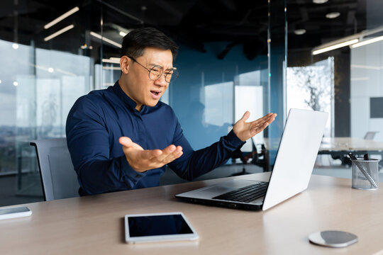 Angry and upset asian businessman, man working with laptop unhappy with result achievement, man inside office disappointed.