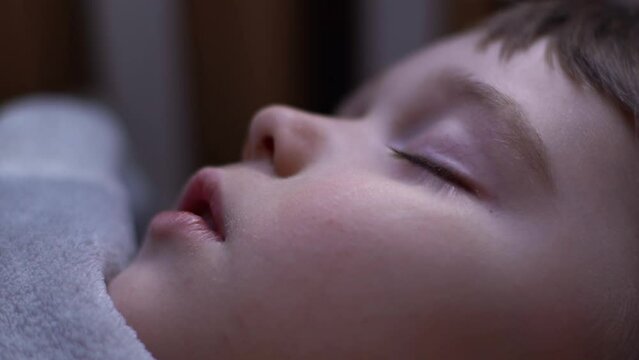 Extreme close-up face of 3-year-old kid boy sleeping with open mouth, breathing deeply, having dream lying down on back under the blanket on bed, in cozy dark children room. Shooting in slow motion.