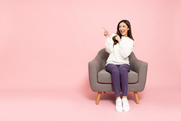 Young elegant Asian woman smiling and pointing to empty copy space and sitting on armchair isolated...