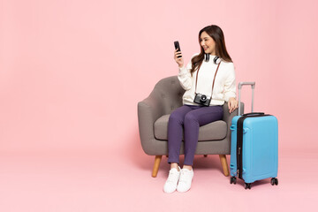 Happy Asian woman traveler sitting on armchair with suitcase and using mobile phone isolated on...