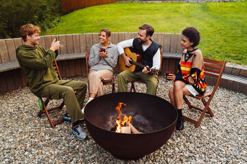 Group of cheerful friends resting by the bonfire, playing the guitar and taking pictures