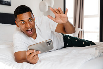 Excited african man playing game on smartphone while lying in bed