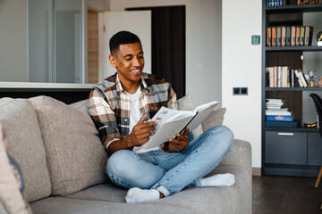 Obraz premium Young smiling african man reading book while sitting on couch at home