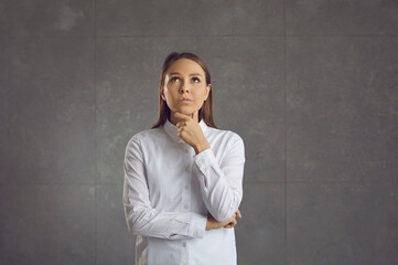 Young caucasian pensive woman thinking and touching chin looking up standing over grey wall...