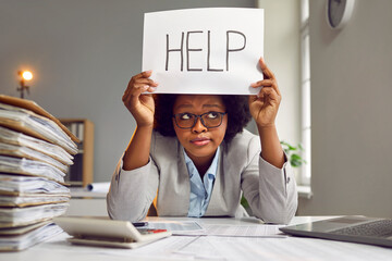 Fototapeta Tired, busy, sad African American woman in a suit and glasses sitting at an office desk with a load of paperwork and holding a paper sign with the word HELP. Working in the office, workload concept obraz