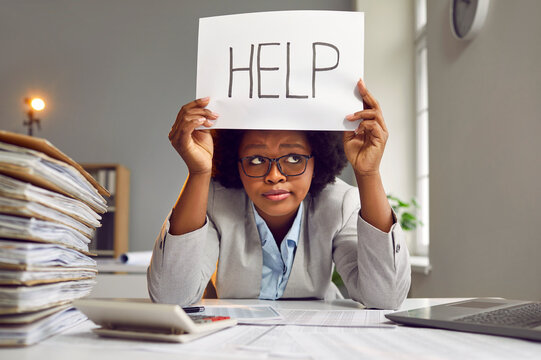 Tired, busy, sad African American woman in a suit and glasses sitting at an office desk with a load of paperwork and holding a paper sign with the word HELP. Working in the office, workload concept