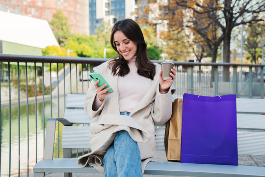 Happy young woman sitting on a bech using a smartphone to text messages after shopping on-line. Brunette female looking to the cellfone device sharing content on social media. High quality photo
