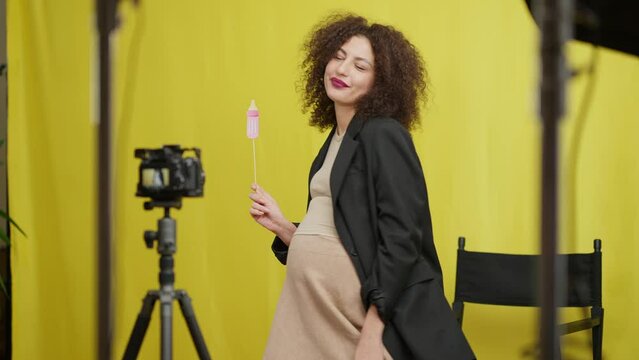 Happy pregnant vlogger talking standing with paper baby pacifier at yellow background recording video blog. Portrait of positive confident Caucasian young beautiful woman filming indoors