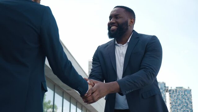 Two smiling male office workers in business suits greet each other with handshake. African american office worker greets male colleague with handshake in downtown. Concept greeting of two business men