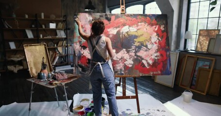 Back view of young woman artist with brush paints on easel with canvas emotional and sensual abstract painting with deep meaning. Female artist makes art in an aesthetic spacious studio. Modern artist - Powered by Adobe