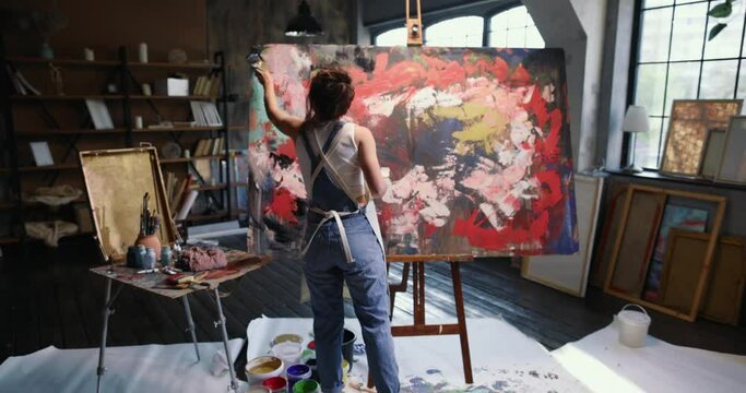 Back view of young woman artist with brush paints on easel with canvas emotional and sensual abstract painting with deep meaning. Female artist makes art in an aesthetic spacious studio. Modern artist