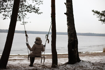 elderly senior retired woman in the forest, background view of a woman from the back on a swing tied to a tree against the backdrop of a lake or river, in winter, unrecognizable