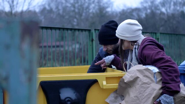 Two dirty and poorly dressed homeless people, a man and a woman, are looking for something in a rubbish bin.