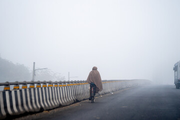 Indian man riding on bicycle in the cold winter morning with dense fog wrapped up tightly showing the harsh winters of Delhi, Rajasthan and Haryana