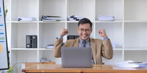 Happy young businessman in suit looking at laptop excited by good news online, lucky successful winner man standing at office desk raising hand in yes gesture celebrating business success win result
