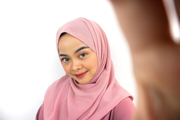 Smiling young Asian Muslim woman dressed in pink doing selfie shot on mobile phone isolated on white background