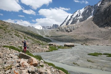 Fototapeta premium Silhouette of woman standing on rock by lake at foot of glacier. Scenery of Tian Shan Mountains during trekking in Altyn Arashan Valley to glacier. Karakol National Park. Kyrgyzstan, Central Asia