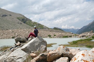 Silhouette of woman sitting on rock by lake at foot of glacier. Scenery of Tian Shan Mountains during trekking in Altyn Arashan Valley to glacier. Karakol National Park. Kyrgyzstan, Central Asia