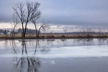 Along a frozen river with melt water, bare trees, and farm fields on a stormy winter day.