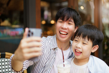 Happy Asian father and son enjoy a selfie photography or video call with their family and friends together while traveling. Cheerful young boy taking and taking a photo on smartphone with his dad.