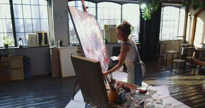 A young modern talented woman artist works on a dark abstract painting with short dotted strokes with a wide brush. Female millennial artist creates modern masterpiece of the oil abstract painting.