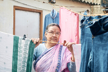 Portrait of a simple looking mature Indian woman (bengali ethnicity) hanging freshly washed laundry to dry in the sun in a winter morning, in building rooftop.