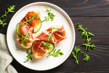 Open sandwiches with cream cheese, prosciutto, cucumber and arugula at wooden table.