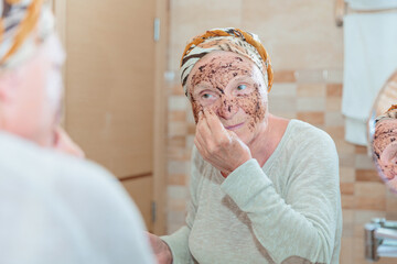 Mature woman standing at the mirror in the bathroom and putting a scrub on her face. Elderly woman taking care of her face