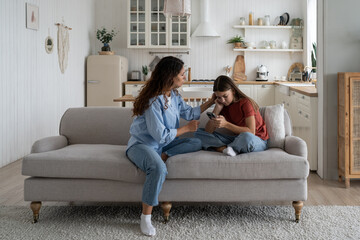 Loving mother sitting with sad daughter on sofa at home, helping child to cope with friendship breakup. Disappointed teen girl looking at phone screen, sharing feelings with mom. First love concept - Powered by Adobe