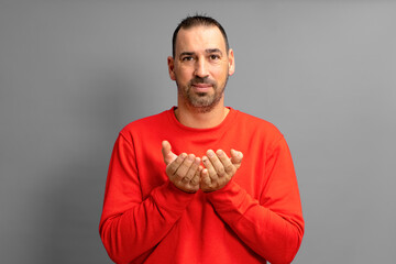 Latino man with beard dressed in a red jumper offering something imaginary in his hands close to the chest isolated on gray studio background, advertising concept, copy space