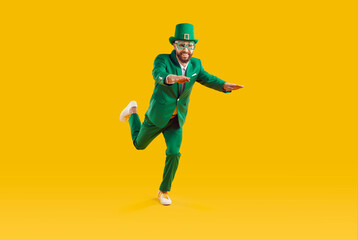Happy man in green carnival costume runs with his arms outstretched in front of yellow background. Isolated object. Running positive leprechaun wearing in clover glasses on bearded face.