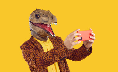 Eccentric funny man in rubber dinosaur mask taking selfie photo on smartphone on yellow background. He wearing leopard jacket and yellow t-shirt. Banner for advertisement, marketing with funky man. - obrazy, fototapety, plakaty