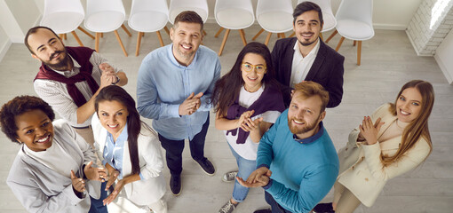 Diverse business team clapping hands. Group portrait of young multiracial people standing together in modern office, looking up at camera, smiling and applauding. High angle, shot from above