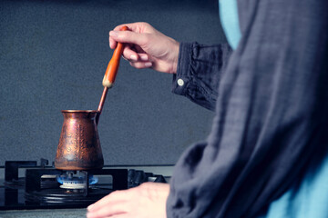 The process of making coffee in an old copper cezve on a black modern stove, a female figure in a gray cotton dress and a linen emerald apron is blurry nearby. Morning coffee ritual Selective focus
