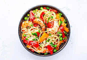Stir fry noodles with shrimps, red and yellow paprika, green pea, chives and sesame seeds in ceramic bowl. Asian cuisine dish. White stone kitchen table background, top view