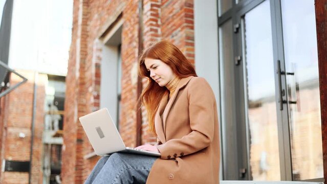 Charming young red haired ginger woman working or studying on laptop sitting on stairs in the urban brick space Busy woman have distance work or online education and looking at the screen outdoors